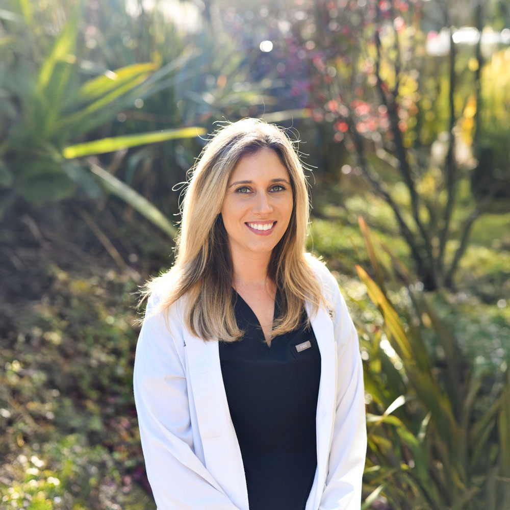 A woman wearing a white lab coat stands outdoors with a bright smile, posing for a professional portrait.